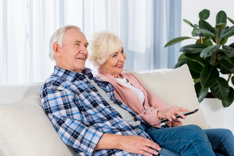 An elderly couple is sitting together on a couch, smiling and looking at something.