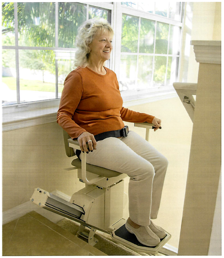A woman smiling while using a stairlift installed in a home with bright windows in the background.