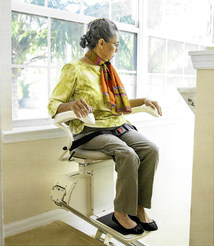 An elderly woman sitting on a stairlift, holding a cup, looking downwards as if reading something.