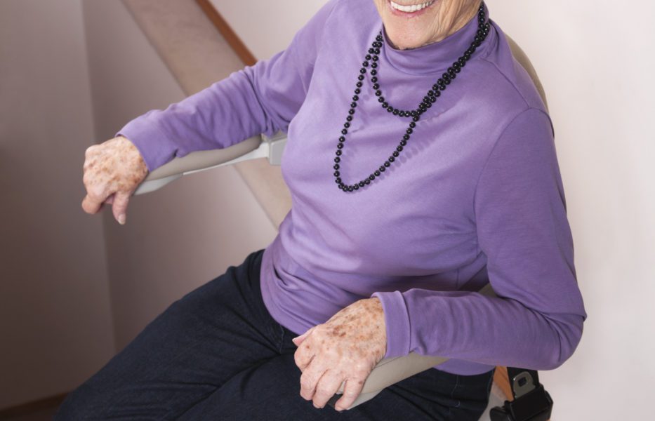 An elderly woman smiles while sitting on a stairlift.