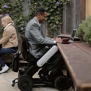 Man in a motorized wheelchair working on a laptop at an outdoor table.