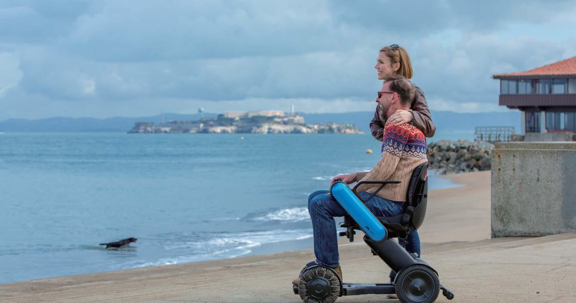 Woman standing behind a man in a wheelchair, both gazing toward the ocean near a coastal promenade.