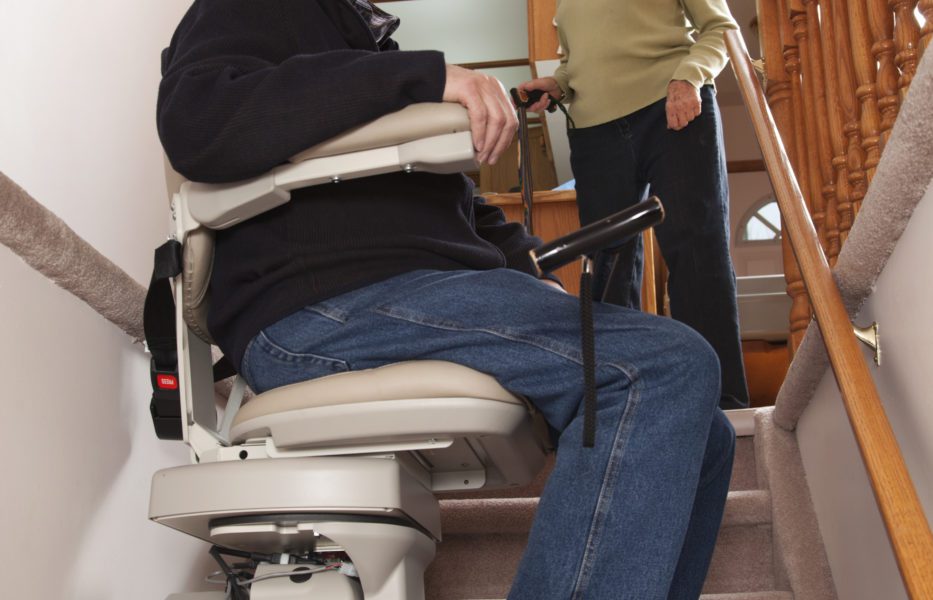 A person using a stairlift to ascend a staircase while another person watches.