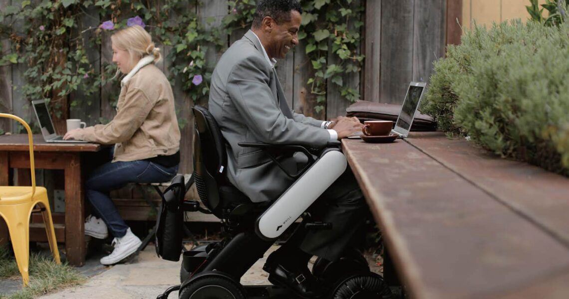 Two people working 2 people working on laptops outdoors; one man in a wheelchair, smiling